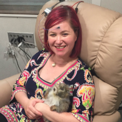 Picture of Dr. Erika Skoe, seated, with sensors placed on her forehead, right ear, and top of head. She is holding the lab mascot, Annabel, a stuffed plushy groundhog.
